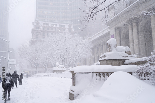 Fotografie pedestrians walking through heavy snowfall beside a historic library plaza with snow-covered trees, statues and columns in a downtown city setting