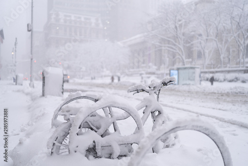 Картината върху платно a bicycle buried in snow on an urban street during heavy snowfall with low visibility, bare trees and buildings in the background