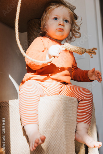 Curious baby sitting on a cat tree and playing with a hanging rope at home. Playful childhood moment in a cozy interior with warm natural sunlight
