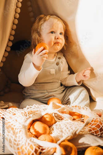 Cute toddler sitting inside a cozy blanket fort and exploring fresh oranges from a reusable mesh shopping bag at home. Candid childhood lifestyle scene with healthy food and imaginative play.
