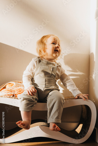 Happy toddler sitting barefoot on a modern chair in a bright sunlit room at home. Candid childhood lifestyle moment with natural light, joyful expression and copy space.