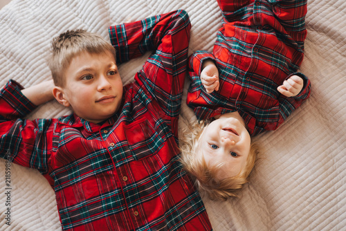 Two brothers lying together on a bed in matching red plaid shirts. Older boy and baby sibling sharing a warm family moment at home, representing brotherhood, childhood and family bonding