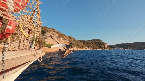 View of a wooden boat navigating near rocky shore and green hills during sunset on a calm sea