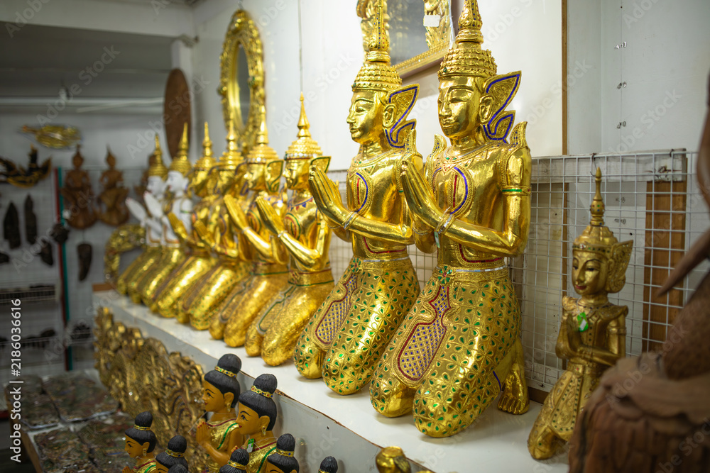 Fototapeta premium Large golden Thepphanom statues stand together with palms pressed in wai prayer pose, traditional Thai guardian figures display intricate craftsmanship inside decorative art shop