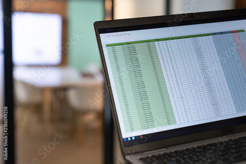 Laptop showing spreadsheet with green column in right foreground, conference room blurring