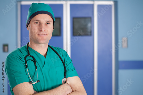 Portrait of confident male surgeon standing with crossed arms and stethoscope in hospital corridor.