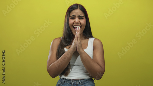 Quadro su tela Young woman wearing white tank top and denim jeans, palms pressed together pleading in studio with yellow backdrop; hopeful anticipation