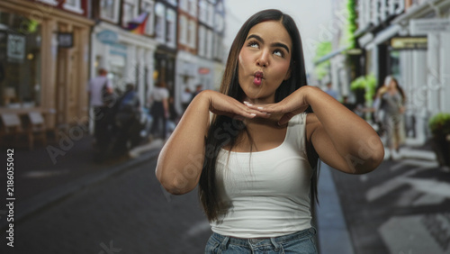 Foto Young hispanic woman smiles showing teeth and bare shoulders on a busy street while making playful hand gestures; joyful