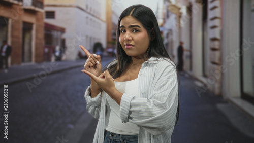 Fotografie Young hispanic woman pointing with index finger and counting gesture on narrow cobblestone street; uncertainty and deliberation