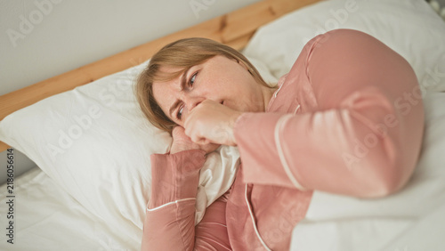 Woman coughing with hand to mouth on bed in bedroom, grimacing while lying on pillow; discomfort illness concern.