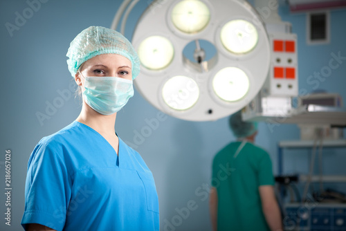Female surgical nurse wearing mask in operating room with surgical lamp and colleague in background.