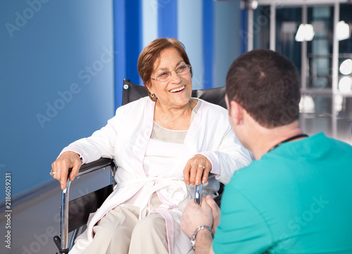 Smiling senior female patient in wheelchair talking with male nurse or doctor in hospital corridor or nursing home.