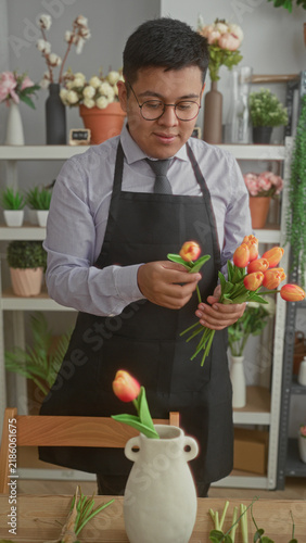 Canvastavla Man placing orange tulips in ceramic vase in studio while wearing apron and glasses, hands arranging flowers; calm care