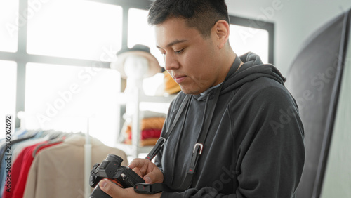 Fotografía Man photographer checking camera touchscreen and adjusting settings in studio with clothing rack, mannequin and softbox visible; concentration