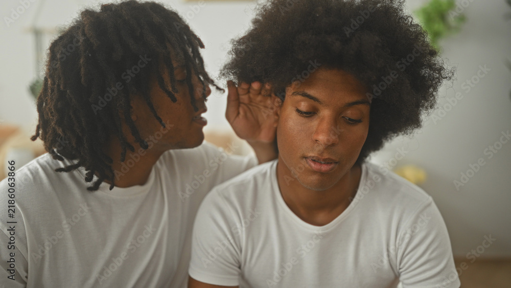 Fototapeta premium Two men on a bed, one whispering into the other's ear while the listener covers his mouth with his hand in a building bedroom setting; quiet intimacy.