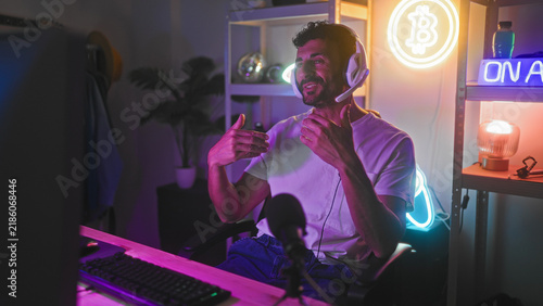 Foto Man wearing headset gesturing hands toward monitor and pointing while seated at computer in a streaming studio with neon signs and mic; engagement connection