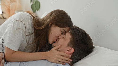 Man lies on bed pillow looking up as woman leans over smiling and touches his shoulder and face in a bedroom inside a building; affection warmth.