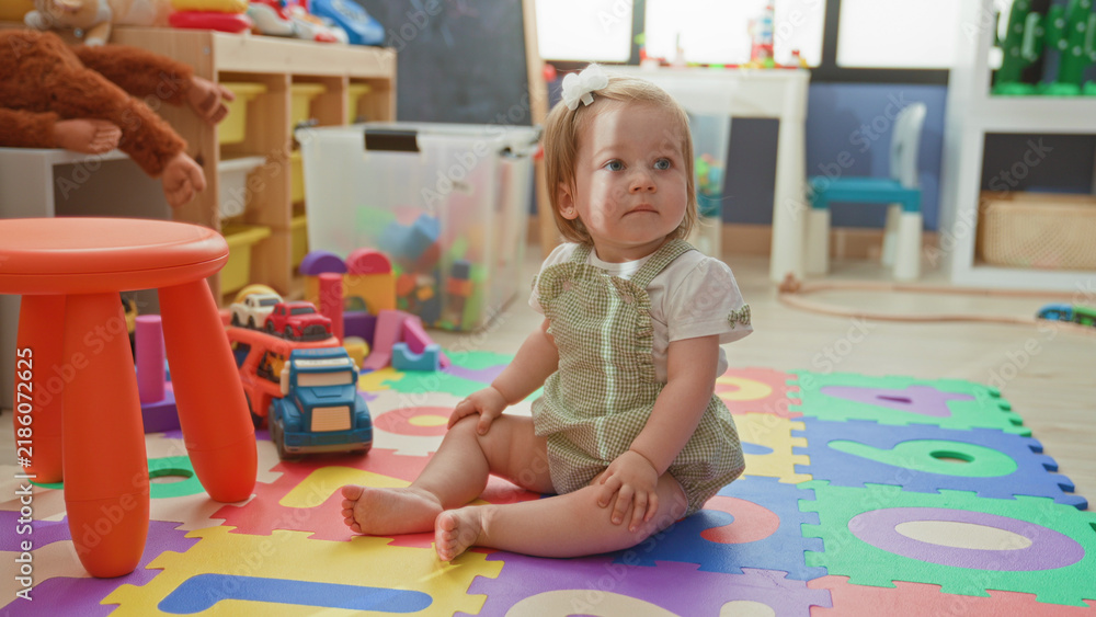 Obraz premium Toddler girl sitting on foam alphabet mat with bare feet and toy truck, looking up in a bright playroom; curiosity innocence.