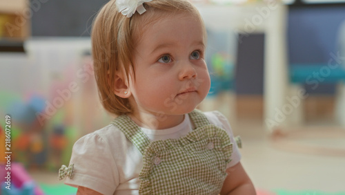Quadro su tela Toddler girl looking up, visible face and white hairbow while seated near colorful toys in a building; innocent curiosity