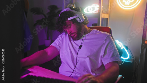 Foto Young hispanic man with beard and curly hair adjusts headset earcup while gripping mouse and leaning forward at a neon lit gaming studio desk; concentration competitive play