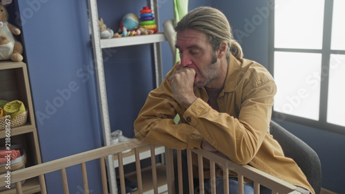 Quadro su tela Man with blond hair rubbing face with hand beside wooden crib and toys on shelf in nursery building; parental exhaustion