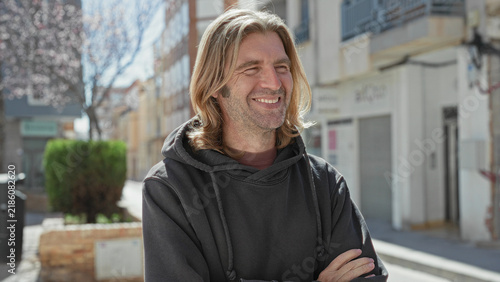Картината върху платно Man in a dark hoodie with arms crossed smiling on a sunlit street in front of shops and a blossom tree beside buildings; contentment relaxed optimism