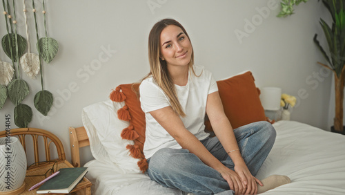 Konstfotografi Woman sitting crosslegged on bed with hands clasped, smiling toward camera, white t shirt and jeans, bedroom; home comfort calm