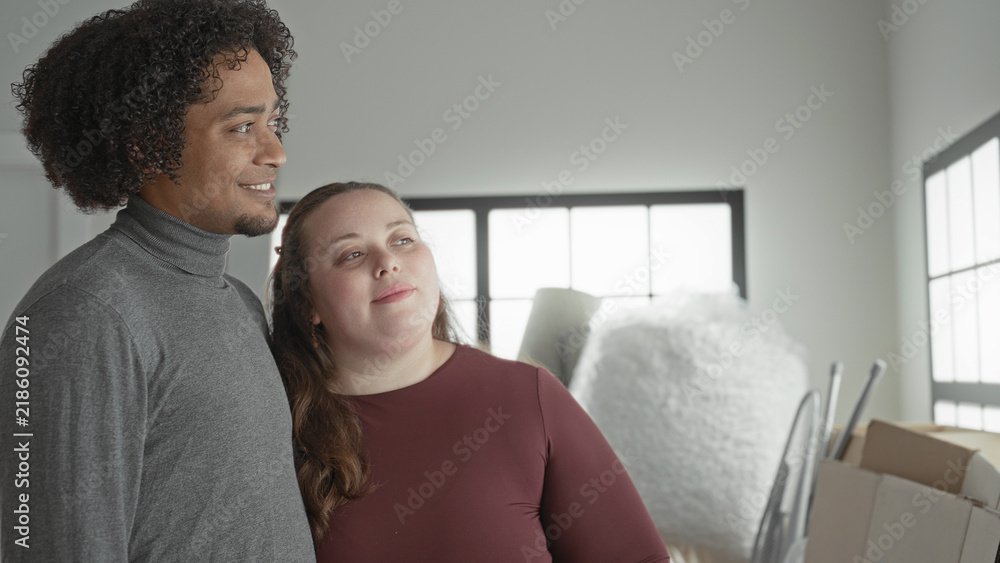 Fototapeta premium Man and plus size woman lean together with head on shoulder, holding moving boxes inside building; affection moving together.