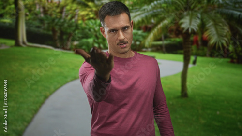 Obraz na plátně Hispanic man with moustache points index finger and beckons toward camera on a forest path wearing maroon long sleeve shirt; assertive command