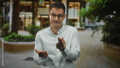 Konstfotografi Man with mustache and glasses in striped shirt holds open palms and gently shrugs in building; uncertainty