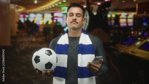 Fotografía Man with mustache wearing a blue and white scarf, holding a soccerball in one hand and a smartphone in the other inside a casino while checking his phone; anticipation camaraderie