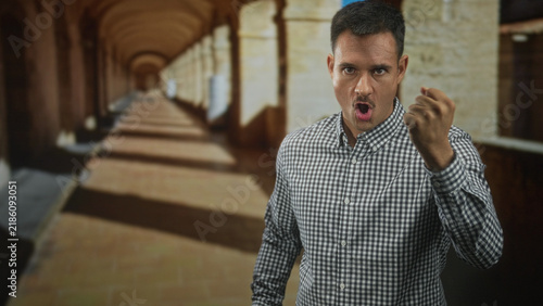 Fotografía Young hispanic man with moustache clenches fist forward in arched building corridor wearing check shirt; anger defiance tension