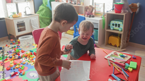 Quadro su tela Two little boys, one crying with mouth open and clutching a crayon while the older brother holds a sticker sheet over a red table in a playroom; sibling conflict frustration