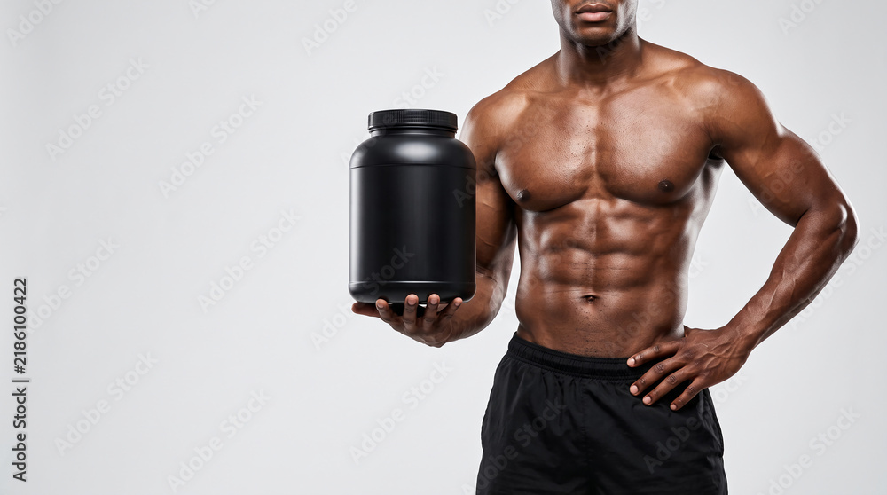 Fototapeta premium Muscular African American man holding a blank black protein powder tub mockup for sports nutrition advertising fit shirtless bodybuilder showing abs against a light gray background with copy space