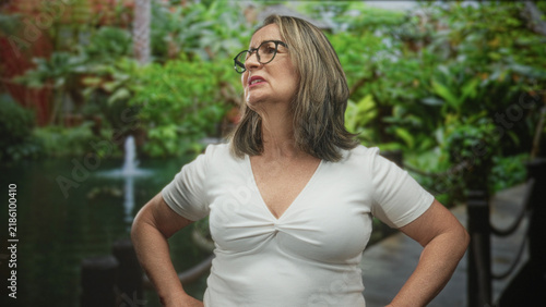 Fotografie Woman with glasses in a white tshirt stands hands on hips by garden pond and fountain, head tilted with pursed lips looking to the side; disapproval