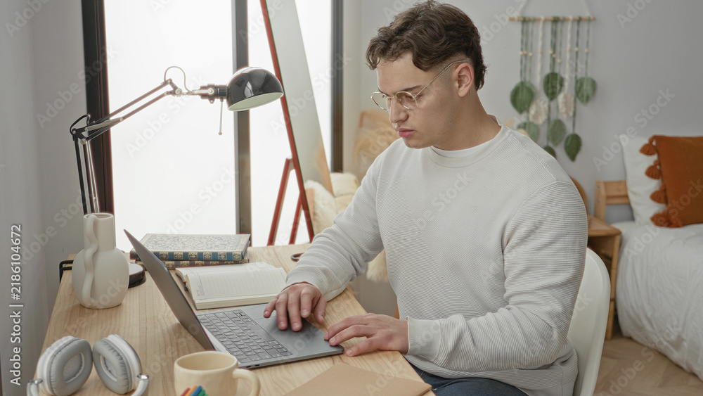 Obraz premium Young man studies at desk, removes glasses and rubs eyes beside laptop in a building; quiet concentration.