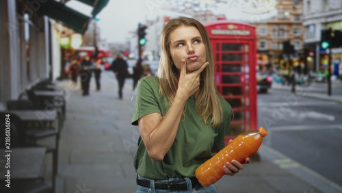 Sticker Blonde woman holding orange juice bottle with hand on chin on street near red phonebox and black taxi; contemplation