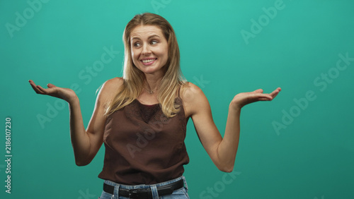 Foto Woman shrugs with palms up and bare forearms visible in a teal studio, blonde young caucasian wearing brown sleeveless top and jeans belt; uncertainty humor