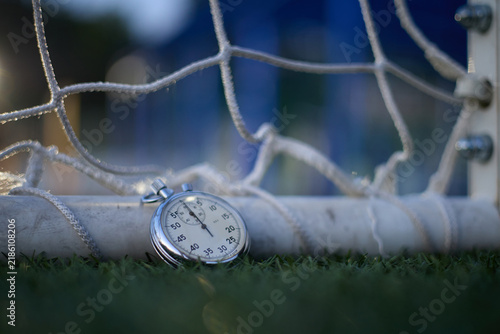 Cinematic close-up of a vintage mechanical stopwatch resting on green grass beside a soccer, with the white net softly blurred in the background. The classic analog dial and precise timing markers