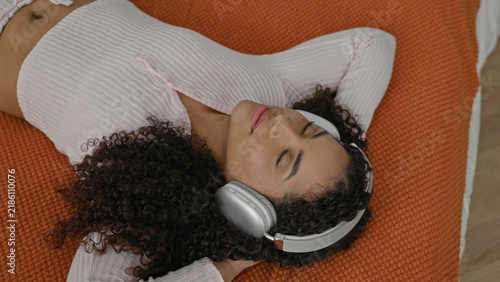 Konstfotografi Woman wearing headphones lying on bed with eyes closed and hand behind head on orange blanket in building; calm relaxation