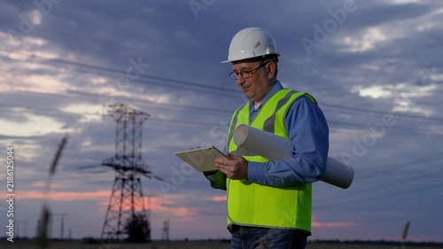 Power engineer in protective white helmet checks power line online using computer tablet, remote access to energy system, for control and safety. High voltage electrical lines at sunset. clean energy