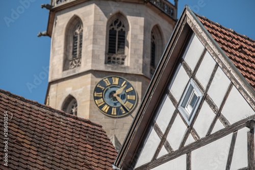 Untersicht Detail Fachwerkhaus mit weißem Kalkputz,  Rautenfachwerk und Rautenfenster vor Kirchturm mit Turmuhr der Stadtkirche in Schorndorf vor blauem Himmel.