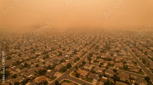 Aerial view of a residential suburb obscured by thick orange smoke haze, illustrating environmental pollution and wildfire impact