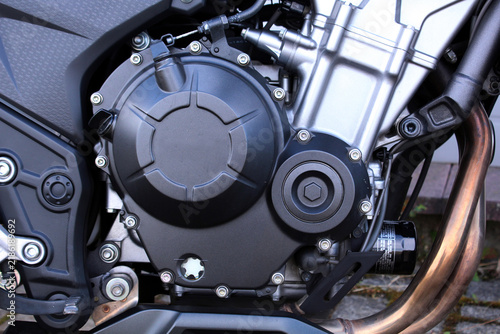 Close-up motorcycle engine with a matte black cover, silver cylinder housing, copper exhaust pipe, circular caps, bolts, hoses, brackets, and detailed mechanical construction