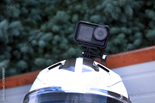 A compact black action camera with a wide lens is mounted on a glossy white motorcycle helmet, featuring curved surfaces, plastic hardware, and a blurred green outdoor background.
