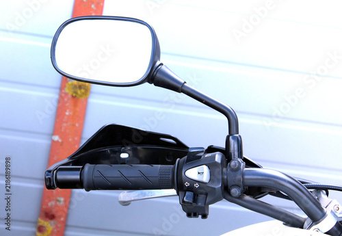 Motorcycle rearview mirror. A black motorcycle handlebar featuring a rearview mirror, a rubber grip, a metal brake lever, and a control unit, set against the backdrop of light-colored garage doors and