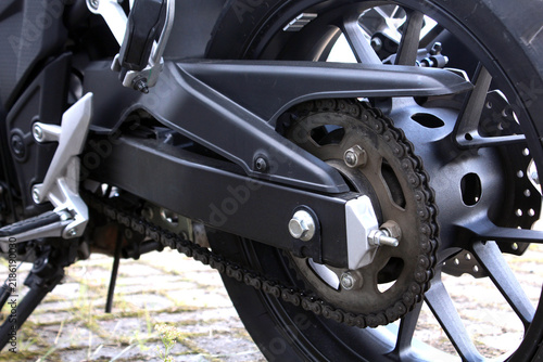 Black motorcycle rear wheel assembly with chain drive, large sprocket, swingarm, alloy rim, silver axle hardware, footrest bracket, brake disc, and rugged mechanical components. Motorcycle chain.