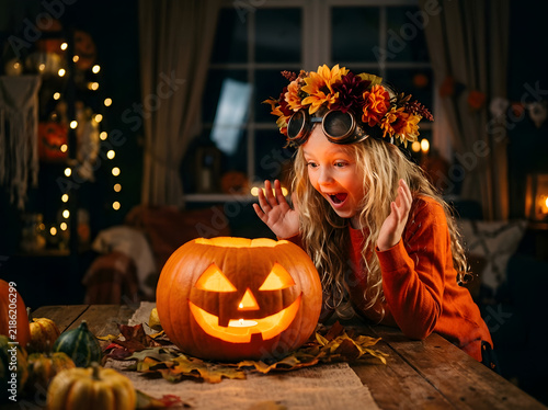 Excited girl with blonde hair wearing floral crown looking at glowing Halloween pumpkin
