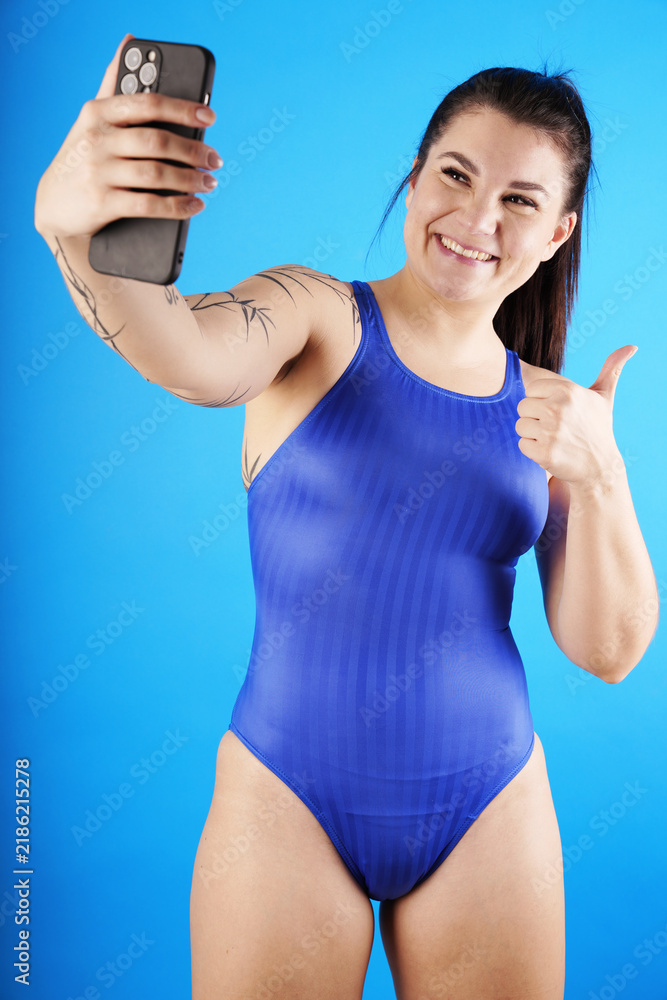 Fototapeta premium A athletic young woman with tattoos wearing a blue shiny one-piece swimsuit is taking selfies with a smartphone camera in studio with blue background