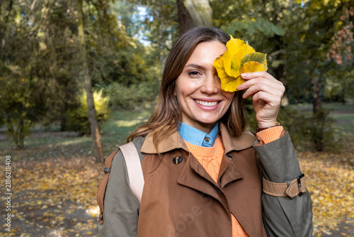 Konstfotografi Woman smiling and holding yellow autumn leaves over her eye in a park